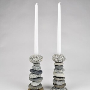 May include: Two candle holders made of stacked stones, each supporting a tall, white candle. The stones vary in size and color, ranging from gray to brown. The candle holders are a natural, rustic home decor item.