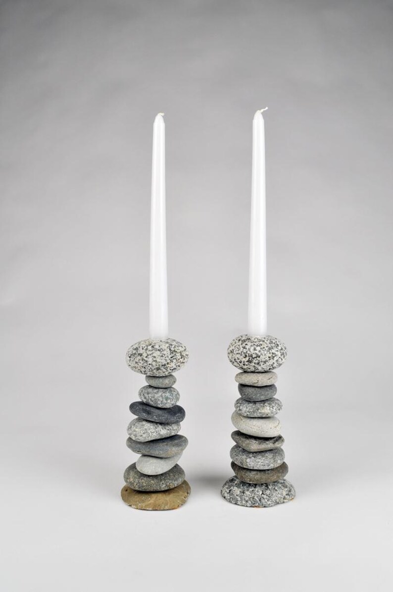 May include: Two candle holders made of stacked stones, each supporting a tall, white candle. The stones vary in size and color, ranging from gray to brown. The candle holders are a natural, rustic home decor item.