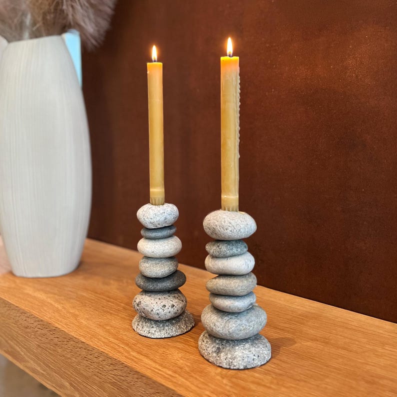 May include: Two lit candles sit atop stacked stone candle holders. The candle holders are made of smooth, gray stones. The candles are a light yellow color and are lit with a small flame. The candle holders are on a wooden surface.
