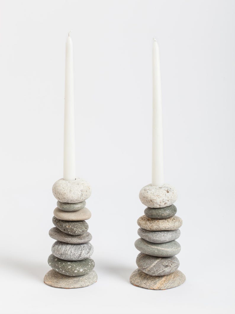 May include: Two candle holders made of stacked, smooth river stones in shades of gray and beige. Each holder supports a tall, white taper candle. The natural stone design adds a rustic, organic touch to home decor.