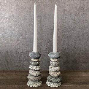 May include: Two stone candle holders with white taper candles. Each holder is made of stacked, smooth, gray and tan river stones. The candles are tall and slender, with a slight curve at the top. The background is a neutral gray.