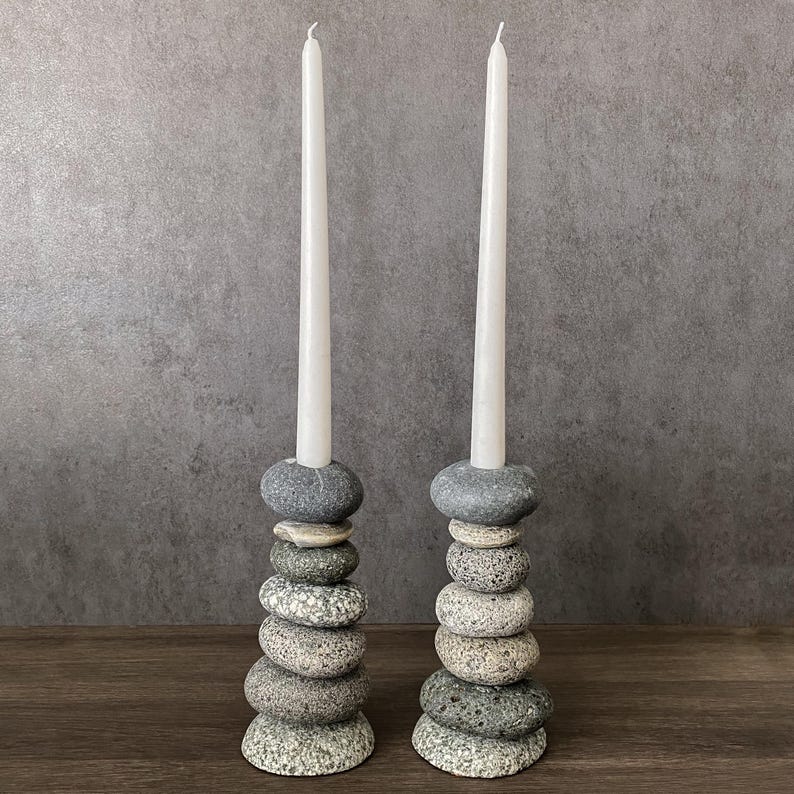 May include: Two stone candle holders with white taper candles. Each holder is made of stacked, smooth, gray and tan river stones. The candles are tall and slender, with a slight curve at the top. The background is a neutral gray.
