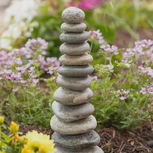 May include: A tall stack of smooth, gray river rocks, carefully balanced on top of each other. The rocks vary in size and shape, creating a natural, zen-like sculpture. The background features a garden with colorful flowers.