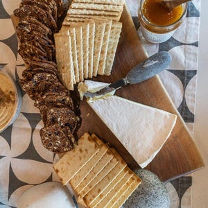 May include: A wooden charcuterie board with crackers, brie cheese, and a jar of preserves. The board also includes a cheese knife, a small candle holder, and decorative stones. The arrangement is set on a patterned cloth.