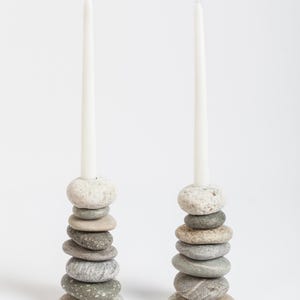 May include: Two candle holders made of stacked, smooth river stones in shades of gray and beige. Each holder supports a tall, white taper candle. The natural stone design adds a rustic, organic touch to home decor.