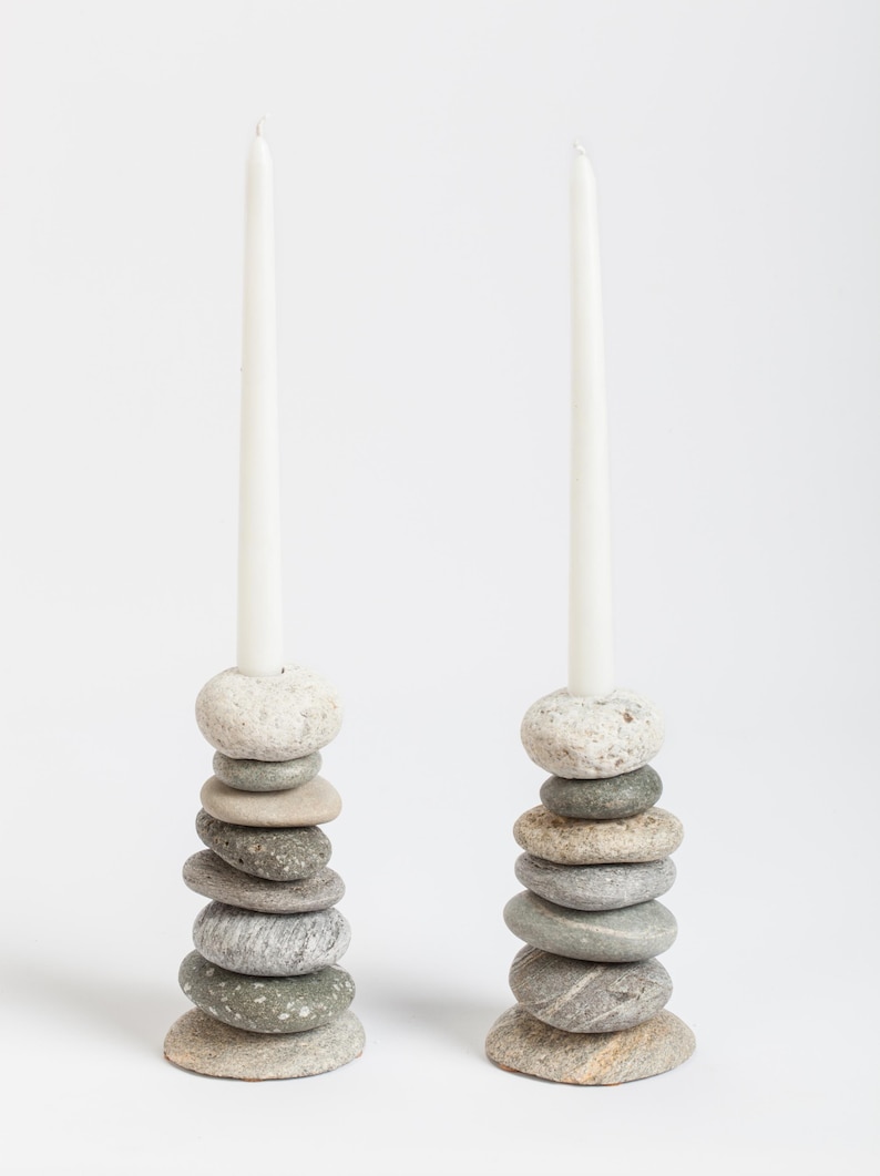 May include: Two candle holders made of stacked, smooth river stones in shades of gray and beige. Each holder supports a tall, white taper candle. The natural stone design adds a rustic, organic touch to home decor.