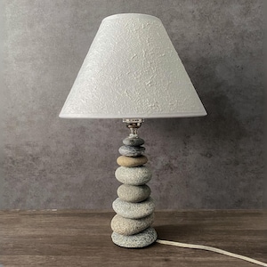 May include: A decorative table lamp with a stacked stone base and a white textured lampshade. The lamp features a silver-colored neck and a neutral color palette, suitable for various interior styles.