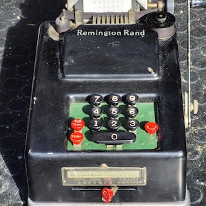 May include: A vintage black Remington Rand mechanical calculator with a paper output. It features black number keys, red total and subtotal buttons, and a crank on the side. The calculator has a green panel with the numbers and a display window.