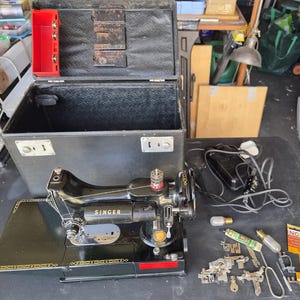 May include: A black Singer sewing machine with gold decorative trim, featuring a matching black carrying case with a red interior compartment, a black foot pedal with a power cord, a small light bulb, and a collection of various metal sewing attachments.