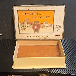 May include: A vintage Rowntree's Chocolates box with a yellow background and a picture of a city scene. The box says "York Assortment" and has a red seal with a white cross.