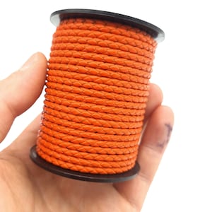 May include: A spool of orange braided cord. The cord is tightly wound around a black plastic spool. The braided texture is visible, and the color is a vibrant orange. The spool is held in a hand against a white background.