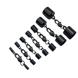 Könnte beinhalten: Ein Set aus vier schwarzen Schmuck-Kordelenden mit befestigten Karabinerverschlüssen, diagonal angeordnet von klein nach groß. Jedes Teil besteht aus zwei zylindrischen Endkappen, die durch eine kurze Kette und einen Karabiner verbunden sind.
