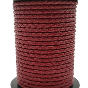 Peut inclure: Une bobine de cordon en cuir tressé de couleur bordeaux. Le cordon est enroulé serré, présentant un motif tissé détaillé. La bobine repose sur une base noire, la couleur riche du cordon contrastant avec le fond blanc.