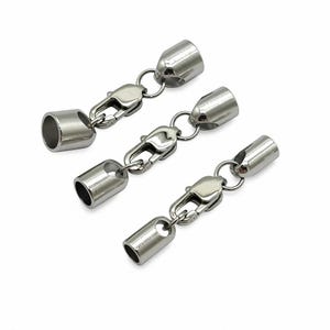 May include: Three silver-toned metal jewelry findings, each consisting of a cylindrical cord end cap connected by a small jump ring to a lobster claw clasp. The components have a smooth, polished finish and are arranged diagonally on a white background.