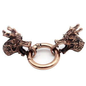 Può includere: Chiusura a testa di drago color bronzo. Due teste di drago dettagliate si fronteggiano, collegate da un anello circolare. La chiusura è in metallo e ha un aspetto invecchiato e antico. Le teste di drago hanno squame intricate e bocche aperte.