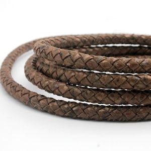 May include: Close-up of brown leather cord, braided in a flat, woven pattern. The cord is soft and supple, with a slightly distressed look.