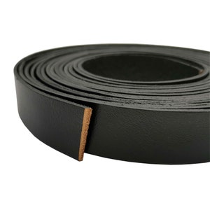 Può includere: Un rotolo di cinturino in pelle nera, largo circa 2,5 cm, con una superficie liscia e testurizzata. La pelle è arrotolata strettamente, mostrando il bordo tagliato della pelle.