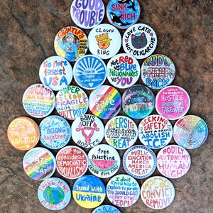 May include: A collection of colorful, round buttons with various hand-drawn designs and text. The buttons feature phrases like "Good Trouble," "Love Cats Not Oligarchs," and "Stand With Ukraine." The overall theme is political activism and social commentary.