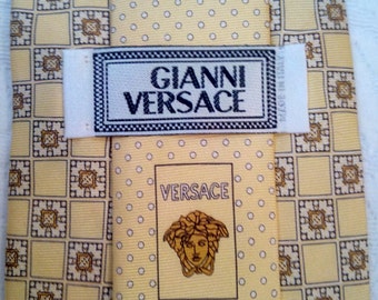 May include: A yellow silk tie with a geometric pattern and a white label that reads "Gianni Versace" and "Made in Italy". The tie also features a smaller label with the word "Versace" and a Medusa head logo.