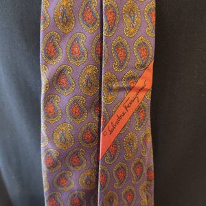 May include: A purple silk tie with a paisley pattern in shades of orange, yellow, and blue. The tie has a red label with the text "Salvatore Ferragamo" in white.