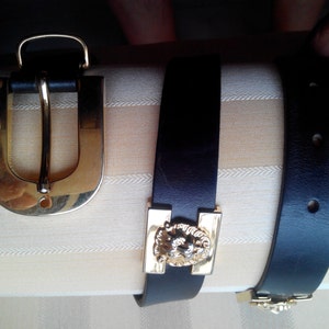 Może przedstawiać: Dwa czarne skórzane paski z pozłacanymi klamrami. Jedna klamra ma prosty prostokątny kształt z motywem lwa. Druga klamra jest duża, w kształcie litery U.