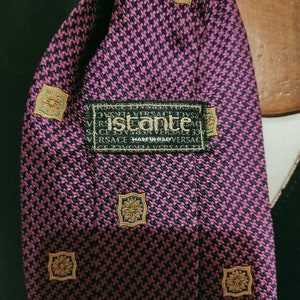May include: A pink and black patterned silk tie with a gold and teal patterned square design. The tie has a label that reads "Istante" and "Made in Italy".