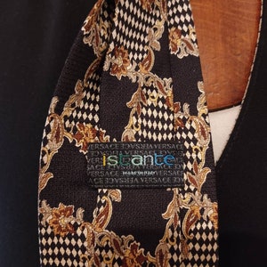 Può includere: Una cravatta di seta nera con un motivo geometrico oro e bianco e accenti floreali. La cravatta ha un'etichetta che recita "ispanto" e "VERSACE MADE IN ITALY".