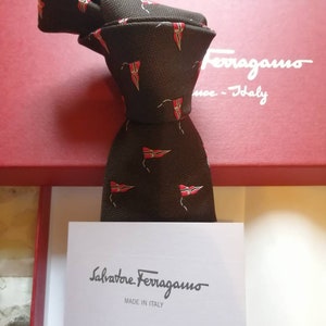 May include: A brown silk tie with a repeating pattern of red and white flags. The tie is in a box with the Salvatore Ferragamo logo and the text "Made in Italy".