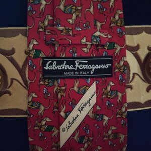 May include: Red silk tie with a repeating pattern of brown dogs wearing saddles and carrying small bags. The tie has a Salvatore Ferragamo label with the text "Salvatore Ferragamo Made in Italy" and a smaller label with the text "Salvatore Ferragamo".