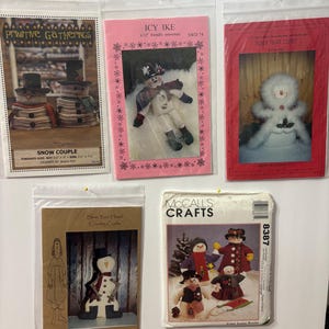 Op de afbeelding: Verschillende naaipatronen met sneeuwmannen, gepresenteerd in transparante plastic hoezen, vastgemaakt op een witte ondergrond. De pakketten hebben titels als "Primitive Gatherings" en "McCall's Crafts". De sneeuwmannen worden in verschillende stijlen afgebeeld.