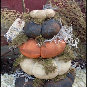 May include: A stacked pumpkin decoration made of fabric in shades of orange, cream, and dark gray, adorned with moss and a tag that reads "Pumpkin Stack". The rustic fall decor is set against a backdrop of hay and wood.