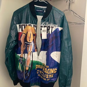 May include: Green and blue Polo Sport bomber jacket with a full-zip closure. The jacket features a cyclist graphic and the text "Le Cyclisme" and "Polo Racing Cyclisme International Tour". The jacket is hanging on a white hanger.