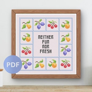 May include: A square cross-stitch pattern framed in wood, featuring a grid of twelve fruit illustrations including cherries, pears, and plums surrounding the black text "NEITHER FUN NOR FRESH" centered on a light pink background with blue borders.