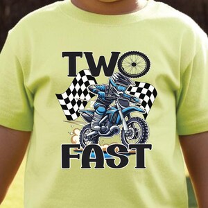May include: A light green t-shirt featuring a motocross design with the text "TWO FAST". The graphic shows a rider on a blue motorcycle with a checkered flag and a bicycle wheel. The shirt is worn by a child.