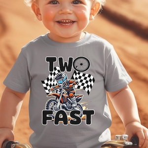 May include: A light gray toddler t-shirt with a motocross-themed graphic. The design features a cartoon rider on a motorcycle, checkered flags, and the words "TWO FAST". The shirt is worn by a smiling toddler.