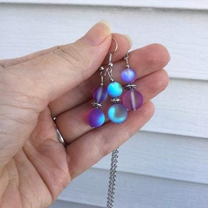 May include: A silver necklace with a iridescent purple and blue bead pendant. The necklace is paired with matching earrings with three iridescent beads each.