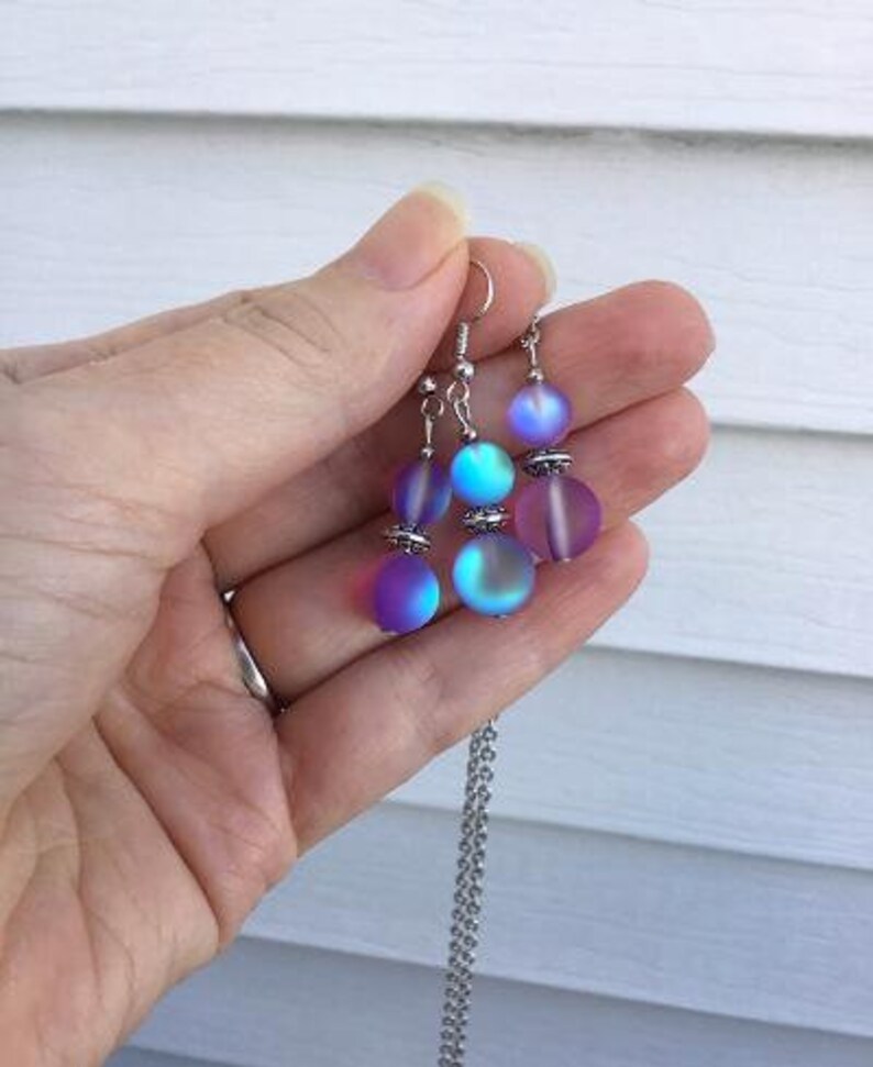 May include: A silver necklace with a iridescent purple and blue bead pendant. The necklace is paired with matching earrings with three iridescent beads each.