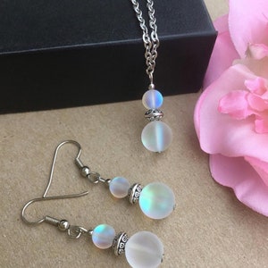 May include: A silver chain necklace with a iridescent white pendant and matching iridescent white earrings. The pendant and earrings are made of two round beads.