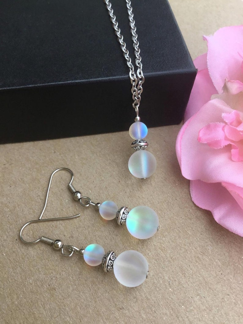 May include: A silver chain necklace with a iridescent white pendant and matching iridescent white earrings. The pendant and earrings are made of two round beads.