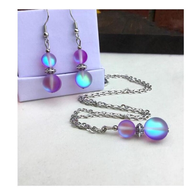 May include: A silver chain necklace with a iridescent purple and blue glass bead pendant. Matching iridescent purple and blue glass bead earrings with silver hooks are also included.