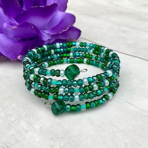May include: A multi-strand wrap bracelet featuring various shades of green and white glass seed beads. The design includes two larger, faceted green beads at the ends of the wire coil, creating a layered appearance with a consistent circular shape.