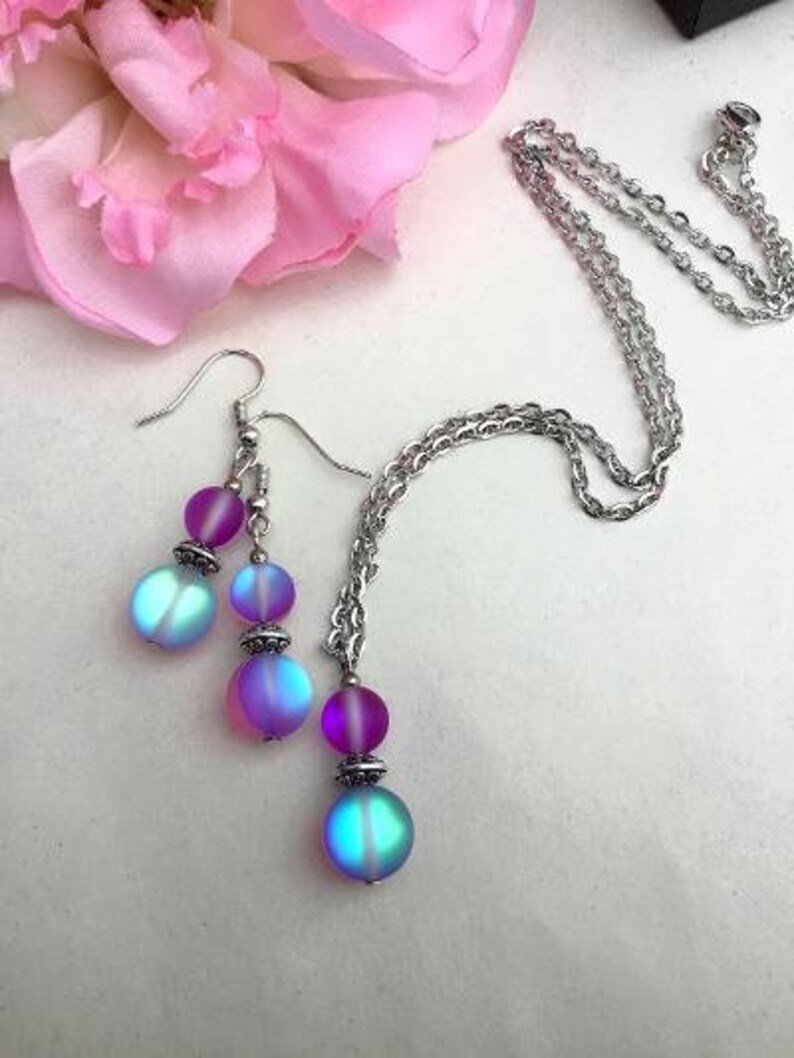 May include: A silver chain necklace with iridescent purple and blue beads. The necklace is paired with matching earrings with iridescent purple and blue beads.