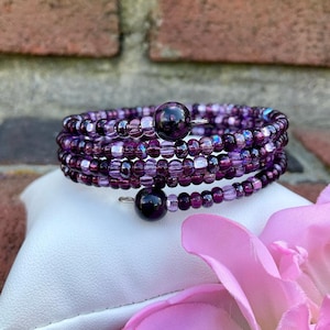 May include: A multi-strand wrap bracelet featuring a variety of small, round beads in shades of deep purple, violet, and light lavender. The bracelet is finished with two larger, dark purple spherical beads at each end of the coiled wire design.