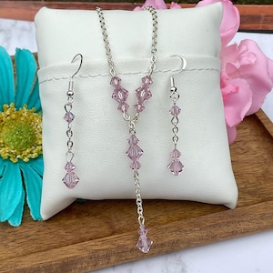 May include: A jewelry set consisting of a silver-toned chain necklace and matching drop earrings. The necklace features a Y-shaped design with light purple faceted crystal beads. Each earring has a silver hook and two light purple crystal beads.