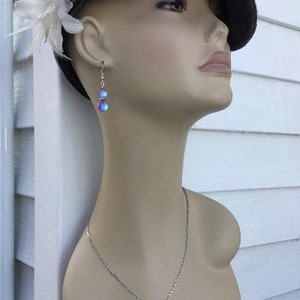 May include: A silver necklace with a pendant featuring two iridescent blue and purple stones. The necklace is worn by a person wearing a gray hat and a white flower.
