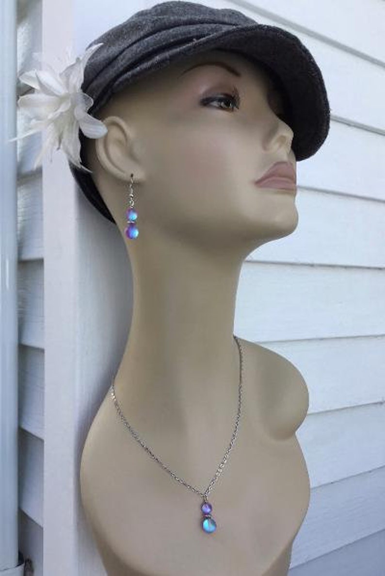 May include: A silver necklace with a pendant featuring two iridescent blue and purple stones. The necklace is worn by a person wearing a gray hat and a white flower.