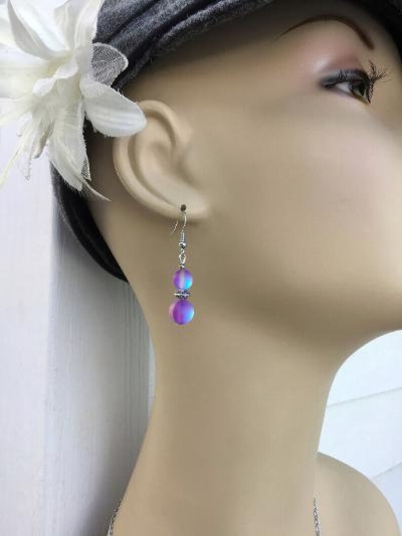 May include: A pair of silver earrings with iridescent purple and blue beads. The beads are round and have a smooth, polished finish.