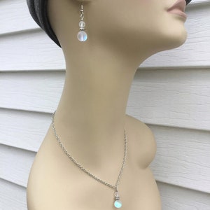 May include: A silver chain necklace with a iridescent white gemstone pendant. The necklace is paired with matching earrings with a smaller iridescent white gemstone.