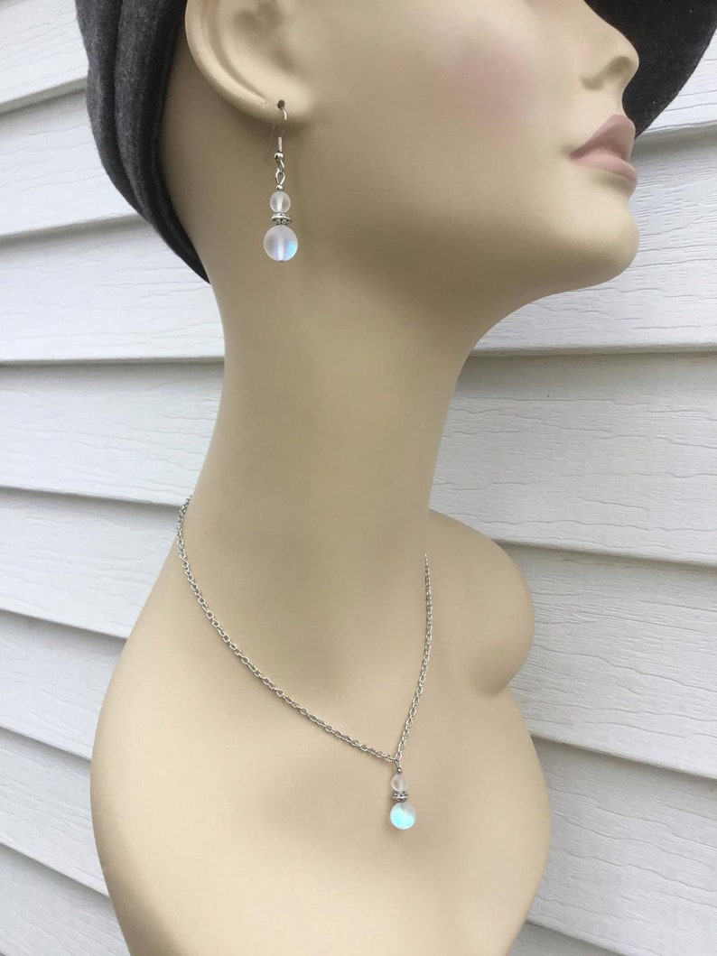 May include: A silver chain necklace with a iridescent white gemstone pendant. The necklace is paired with matching earrings with a smaller iridescent white gemstone.