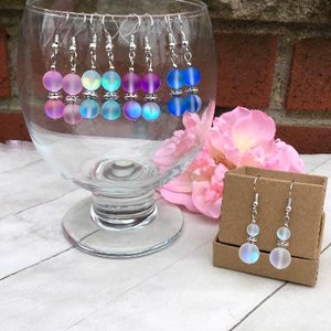May include: A set of iridescent earrings with silver hooks and two round beads. The earrings are displayed in a glass bowl on a white surface.